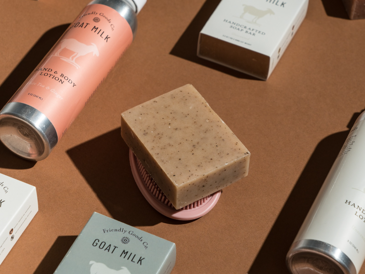 Friendly Goods products displayed in a pattern on a brown background with an unwrapped goat ilk soap bar front and center on a pink silicone soap lift