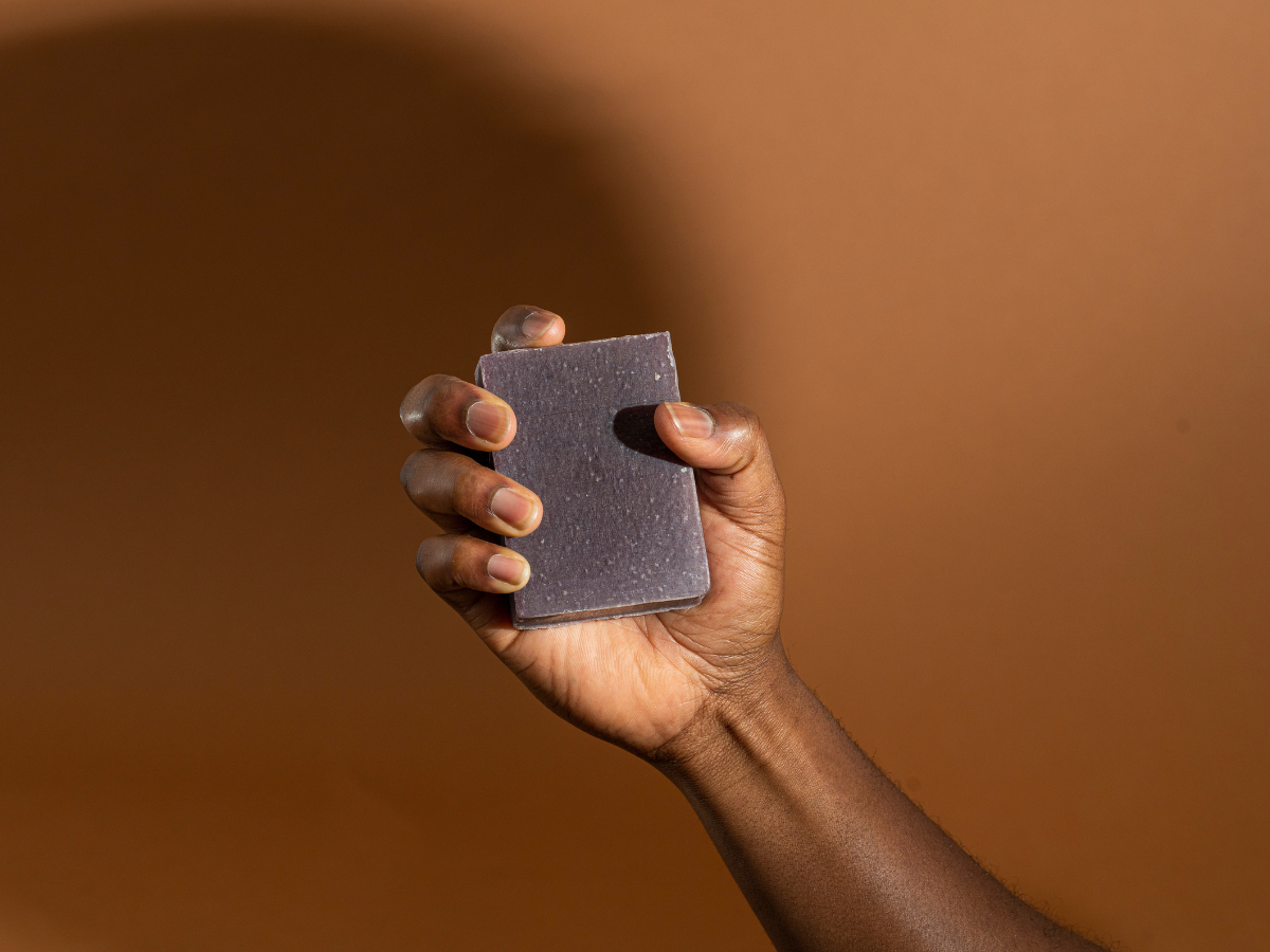 Black male hand gripping a Midshipman Goat Milk soap bar against a brown background