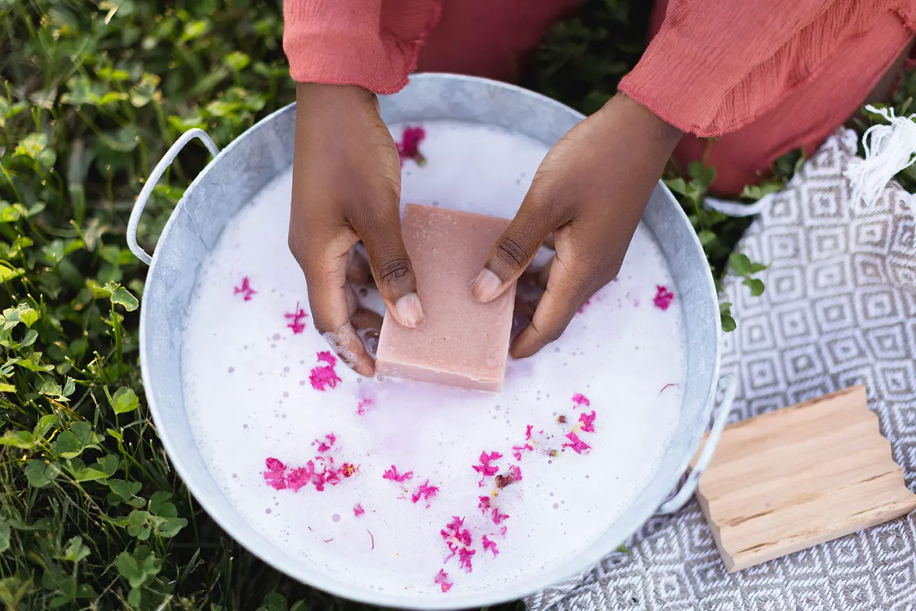 Two hands holding a friendly goods goat milk soap bar in an aluminum wash basin with pink flower petals